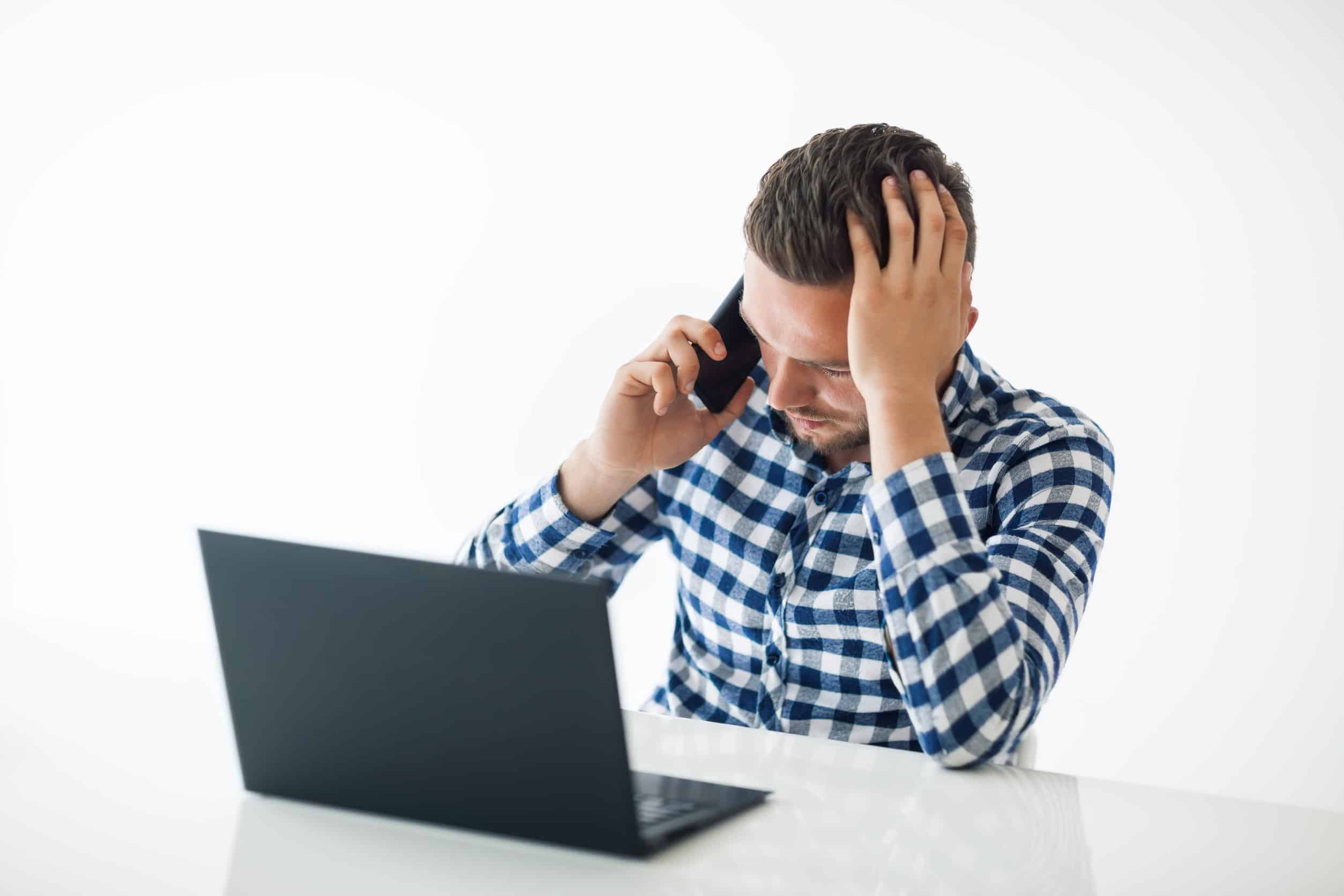 Stressful tired young businessman with laptop speaking on mobile phone at the office on white background